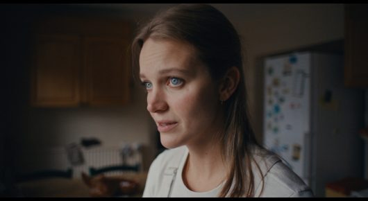 Still from Cambridge Connections: a close-up of a young woman looking to one side in a dim kitchen, with a fridge covered in magnets behind her.