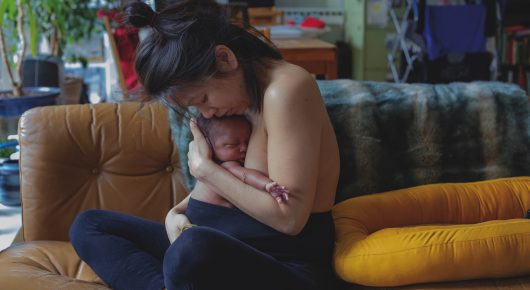 Still from Cambridge Connections: a woman on a sofa holds a baby against her bare chest in a living room, with yellow cushions beside her.