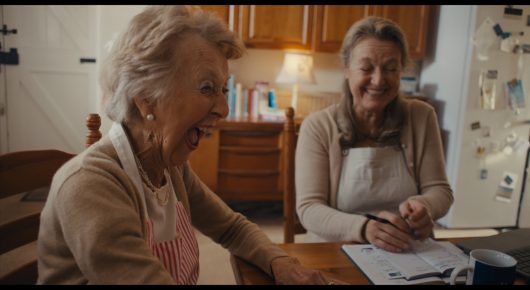 Still from Cambridge Connections: two older women sit at a kitchen table laughing, one in an apron with a notebook in front of her.