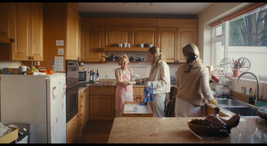 Still from Cambridge Connections: three women work in a wooden kitchen, seen from across the room, one of them at the sink.