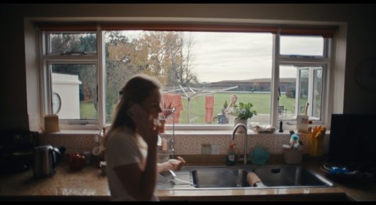 Still from Cambridge Connections: a woman stands by a kitchen window overlooking a field.