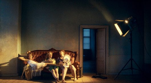 Still from A Sweetness from Nowhere: two people sit on a red sofa in a large, dim room with a tall stage lamp beside them.