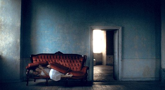 Still from A Sweetness from Nowhere: a person lies on a red sofa in a large, dim room, with an open doorway of bright light behind.