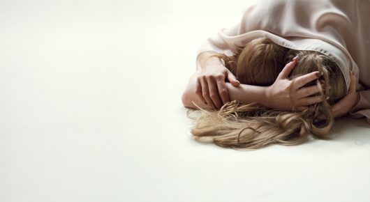 Still from A Sweetness from Nowhere: a person with blonde hair lies curled on a white floor, covering their face.