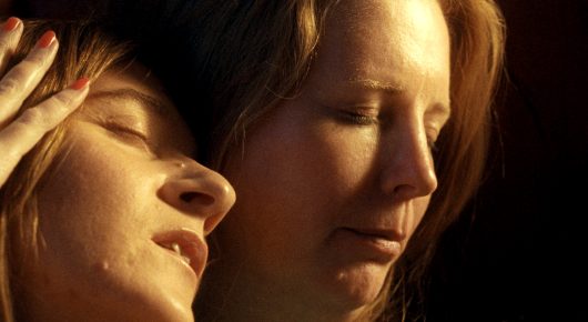 Still from A Sweetness from Nowhere: a close-up of two women with their faces close together, one with her eyes closed, in warm golden light.