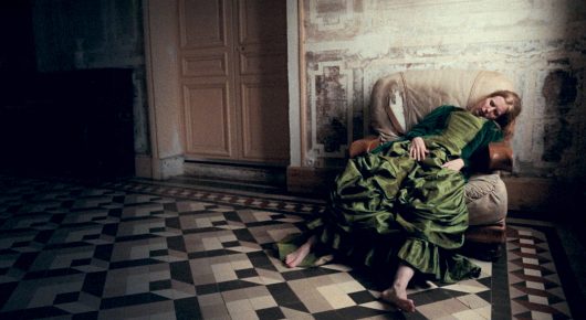 Still from A Sweetness from Nowhere: a woman in a green dress lies on a black-and-white tiled floor in an ornate room.