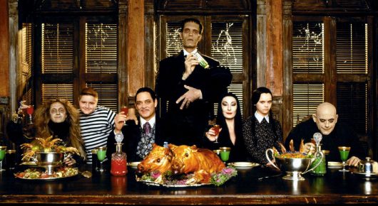 Still from Addams Family Values: the Addams family sit around a long table laden with food in a dark wood-panelled dining room.