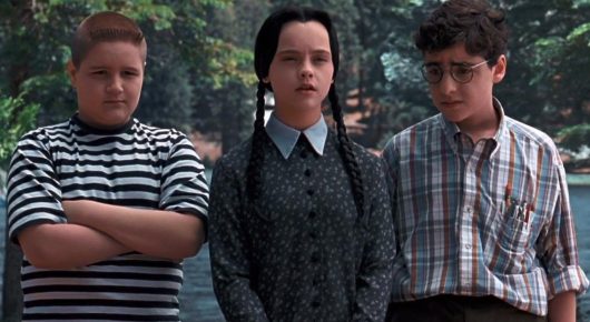 Still from Addams Family Values: two boys and a girl with black braids in a grey dress stand side by side, the boys in striped and checked shirts.
