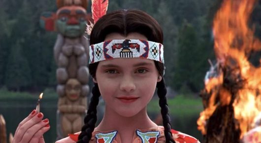 Still from Addams Family Values: a girl with long braids, a feathered headband and face paint smiles, holding a lit match in front of a large bonfire.