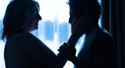 Still from Ali & Ava: two people stand close together in silhouette against a bright window, one with a hand raised to the other's face.