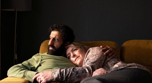 Still from Ali & Ava: a bearded man in a green jumper and a woman lie close together on a yellow sofa in a dim room.