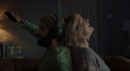 Still from Ali & Ava: a bearded man wearing headphones holds one arm raised while a woman leans back against him with her head tilted up, in a dimly lit room.