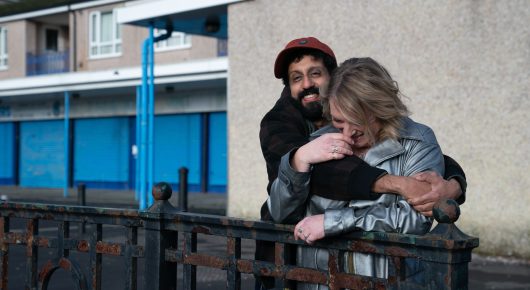 Still from Ali & Ava: a bearded man in a red cap and a woman in a silver jacket hug on a street outside a block of flats.