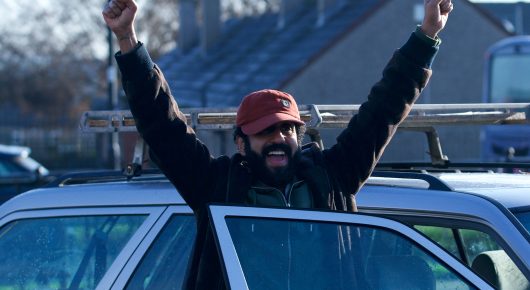 Still from Ali & Ava: a bearded man in a red cap stands by a parked car with both arms raised, with houses behind him.