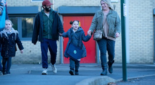 Still from Ali & Ava: a man, a woman and two children walk along a street past a red door, with a girl in a blue coat holding the man's hand.