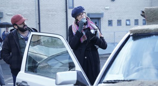 Still from Ali & Ava: a man in a red cap stands by an open car door while a woman in a purple hat and dark coat talks on a phone on the pavement.