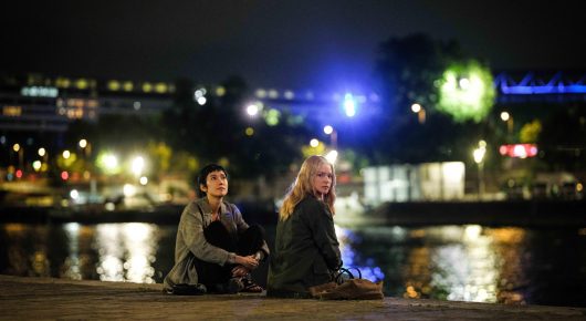Still from All of a Sudden: two people sit on a stone quayside at night, with city lights reflected in the river behind them.
