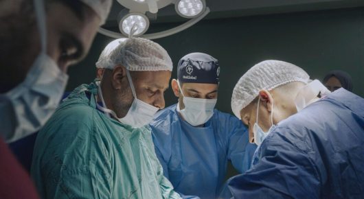 Still from American Doctor: surgeons in scrubs, masks and hair nets work over an operating table under bright lights.