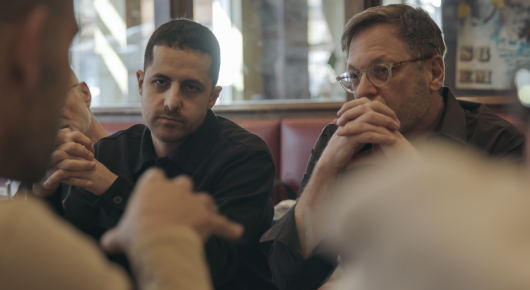 Still from American Doctor: two men sit side by side in a booth with their hands together, one with dark hair and one with glasses and a goatee.