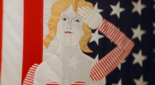 Still from Artists in Residence: an artwork of a woman with yellow hair raising her hand in a salute, set against a red-striped, star-covered flag.