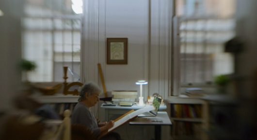 Still from Artists in Residence: a blurred view of a person sitting at a desk with a lamp in a bright, quiet room.