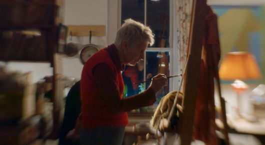 Still from Artists in Residence: an older woman in a red top and apron paints at an easel in a cluttered studio with a lamp.