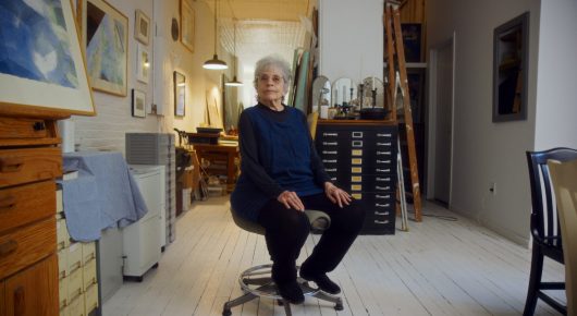 Still from Artists in Residence: an older woman in a navy top sits on a swivel chair in a cluttered studio with shelves and paintings.