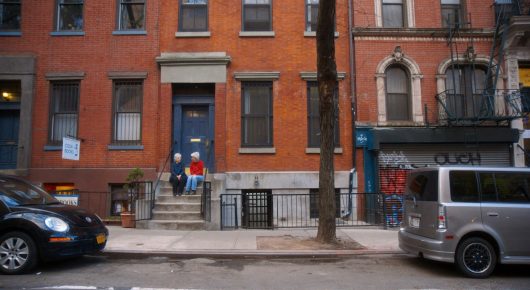 Still from Artists in Residence: a red-brick townhouse street with parked cars and a van, with steps up to a blue front door.