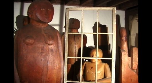 Still from Artists in Residence: wooden figures, including a large carved figure and smaller ones, stand behind a white-framed window panel in a workshop.