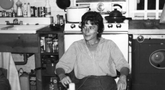 Still from Artists in Residence: a black-and-white photograph of a woman with short dark hair sitting on a chair in a kitchen with an old stove and shelves.