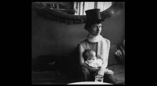 Still from Artists in Residence: a black-and-white photograph of a woman in a top hat holding a baby, seated in front of a curved background.