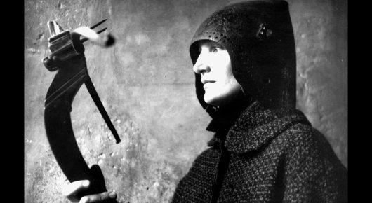 Still from Artists in Residence: a black-and-white photograph of a person in a dark hooded coat in profile, beside a dark curved object.
