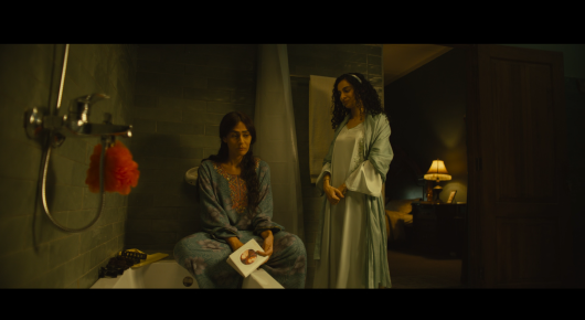 Still from Baab: two women in a dim room, one in a long pale gown and one in a blue top, with a shower head on the wall behind them.