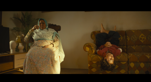 Still from Baab: an older woman in a white headscarf sits in an armchair while a child lies upside down on a patterned sofa.
