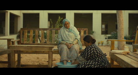 Still from Baab: an older woman in a pale headscarf sits on a bench in a courtyard while another woman kneels in front of her beside a blue basin.