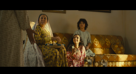 Still from Baab: an older woman in a patterned yellow dress sits on a gold sofa with two children, while another person stands at the edge of the frame.