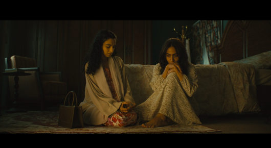 Still from Baab: two girls in pale robes sit on the floor of a dim bedroom, one with her hands raised to her mouth.