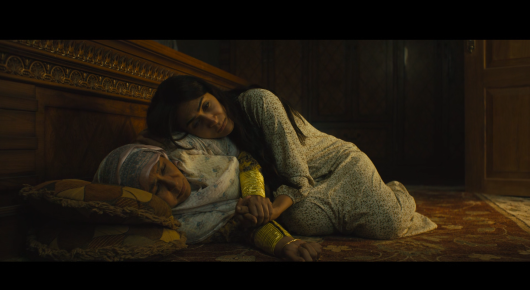 Still from Baab: two people in pale clothes lie close together on a rug and cushions in a dim, wood-panelled room.