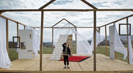 Still from The Beauty of the Donkey: a small figure in dark clothes stands on a wooden platform inside an open timber-framed house with white curtains blowing, under a cloudy sky.