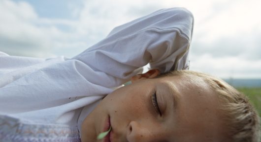 Still from The Beauty of the Donkey: a close-up of a blond child asleep with their head on white fabric and a stalk of grass in their mouth.