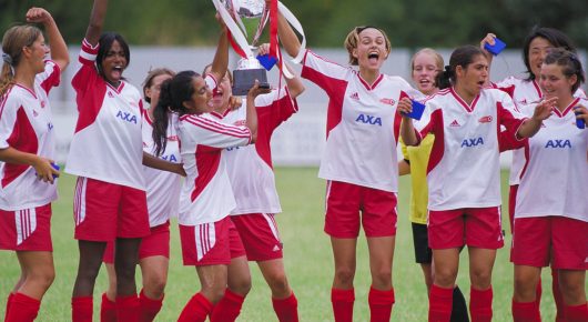 Still from Bend It Like Beckham: a women's football team in red and white kit celebrates on a pitch, one player holding a trophy aloft.