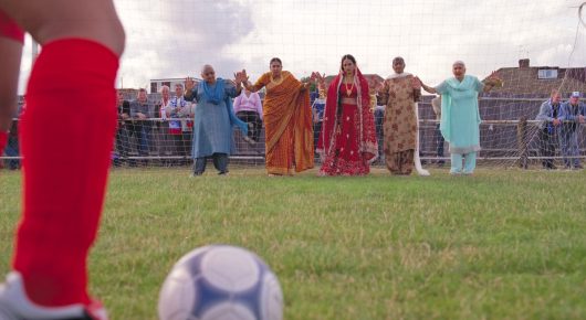 Still from Bend It Like Beckham: a football on grass beside a player's red sock and boot in the foreground, with a group of people in colourful saris standing behind a fence.