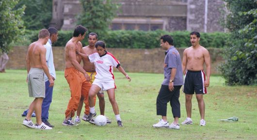 Still from Bend It Like Beckham: a group of men, some shirtless, kick a football around on a grassy park, with a girl in a white top among them.