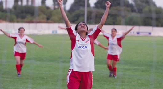 Still from Bend It Like Beckham: a young woman in a red and white kit raises both arms in celebration on a football pitch, with teammates behind her.