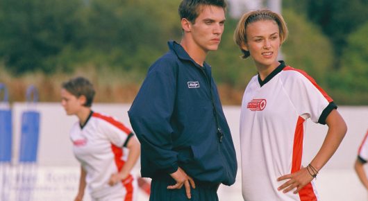 Still from Bend It Like Beckham: a man in a navy jacket and a young woman in football kit stand on a pitch talking, with another player behind them.