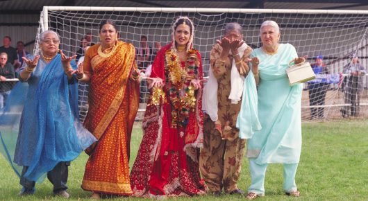 Still from Bend It Like Beckham: five women in brightly coloured saris and salwar kameez stand in front of a football goal on a pitch.