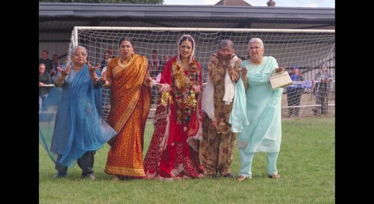 Still from Bend It Like Beckham: five women in brightly coloured saris and salwar kameez stand in front of a football goal on a pitch.
