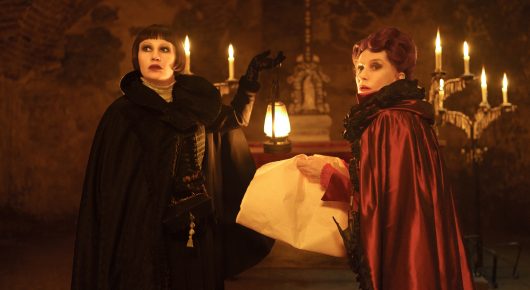 Still from The Blood Countess: two figures in black capes stand by a table of candles, one holding a folded document.