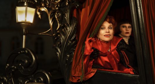 Still from The Blood Countess: two women in red and dark clothes look out from a theatre box with a red curtain and a lantern.