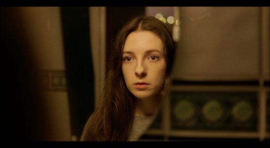 Still from Cambridge Connections: a young woman with long hair looks at the camera in a dim room.
